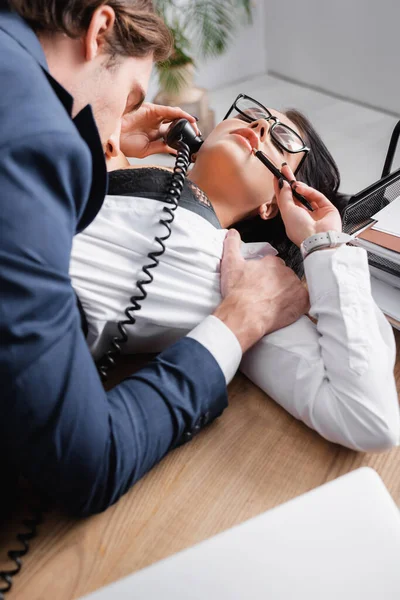 Бизнесмен обнимает сексуальную секретаршу лежа на столе и разговаривая по телефону — стоковое фото