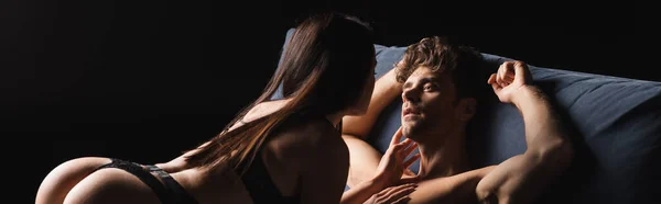 Чуттєва жінка торкається сорочки хлопця на ліжку ізольовано на чорному, банер — стокове фото