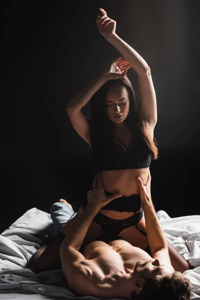 Мужчина без рубашки касается талии чувственной женщины в нижнем белье и поясе подвязки на кровати изолированы на черном — стоковое фото