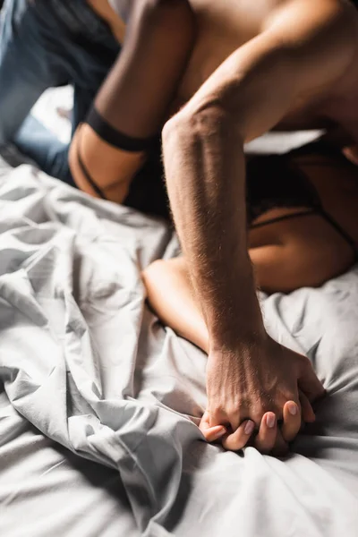 Обрезанный вид сексуальной пары на размытом фоне, держащей руки на белых постельных принадлежностях — стоковое фото
