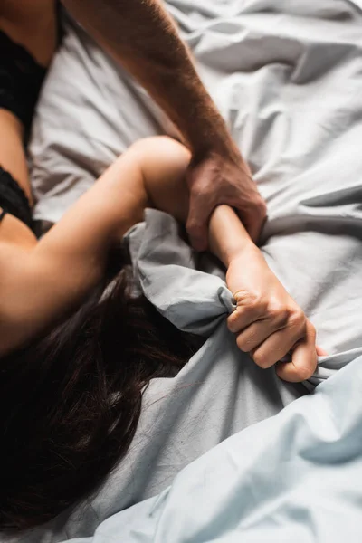 Высокий угол обзора мужчины, держащего за руку размытую девушку в нижнем белье на кровати — стоковое фото