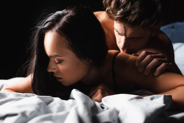 Чувственная женщина, лежащая рядом с расплывчатым бойфрендом на кровати, изолированная от черного — стоковое фото