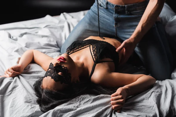 Mulher sensual em máscara de renda deitada na cama perto do namorado com palmada remo no fundo preto — Fotografia de Stock