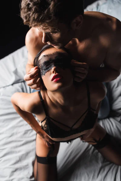 Vista superior do homem sem camisa usando máscara de renda na namorada sensual na cama isolada no preto — Fotografia de Stock