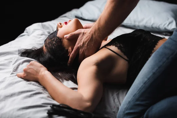 Homme en jeans toucher le cou de femme sexy en soutien-gorge et masque de dentelle sur le lit sur fond noir — Photo de stock