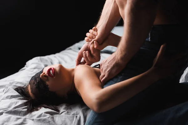 Чоловік торкається пристрасної дівчини в мереживній масці на ліжку ізольовано на чорному — стокове фото