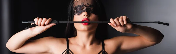 Sensual woman in lace mask and bra holding spanking paddle on black background, banner — Stock Photo