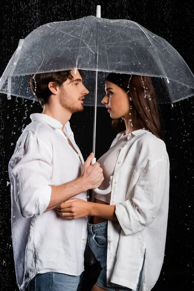 Vista lateral de romántico joven novio adulto y novia de pie en la lluvia con paraguas sobre fondo negro - foto de stock