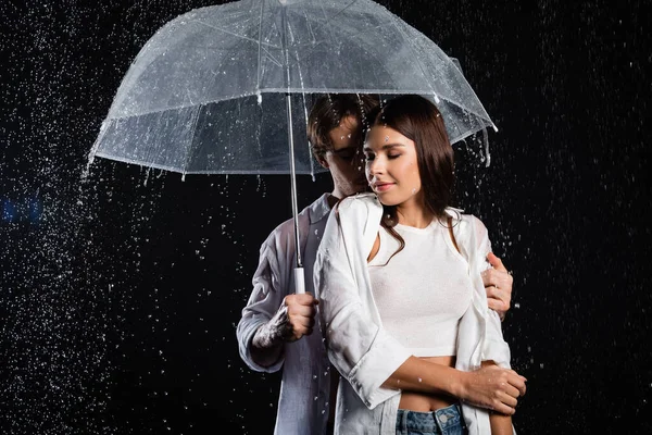 Романтическая молодая пара, стоящая под дождем с зонтиком и чувственно обнимающая на черном фоне — стоковое фото