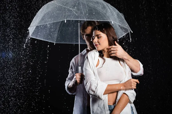 Romantic young adult couple standing in rain with umbrella and sensually hugging on black background — Stock Photo