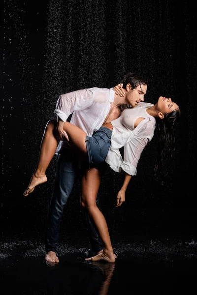 Mojado hombre guapo apasionadamente abrazando a la mujer y sosteniendo su pierna a mano en gotas de lluvia sobre fondo negro - foto de stock