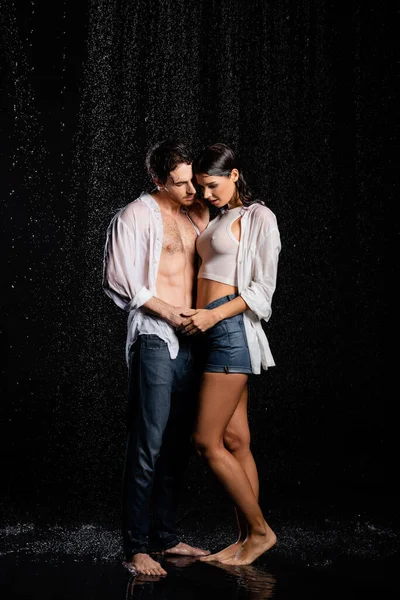 Wet handsome man gently hugging and holding hands with pretty woman in rain drops on black background — Stock Photo