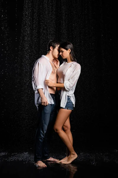 Vista completa de pareja sexy abrazos y besos en ropa mojada en gotas de lluvia sobre fondo negro - foto de stock