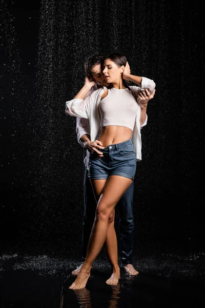 Вид в полный рост сексуальной пары, обнимающейся в мокрой одежде в каплях дождя на черном фоне — стоковое фото