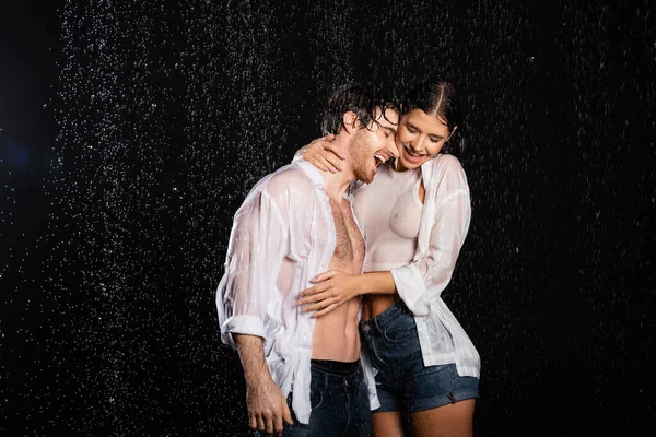 Smiling boyfriend embracing with girlfriend in wet clothes in rain drops on black background — Stock Photo