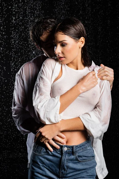 Novio abrazando con novia en mojado ropa en gotas de lluvia en negro fondo - foto de stock