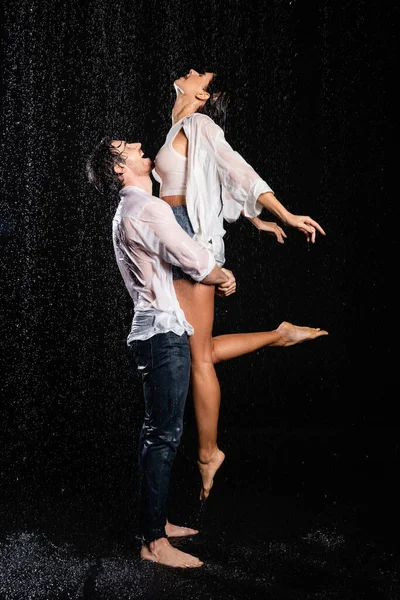 Petit ami heureux levant petite amie souriante en vêtements humides sous la pluie gouttes sur fond noir — Photo de stock