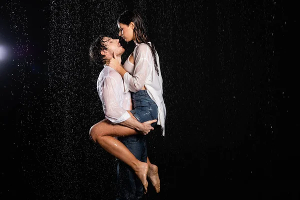 Сексуальная романтическая пара в мокрых белых рубашках и джинсах обнимающая и целующаяся в капли дождя на черном фоне — стоковое фото