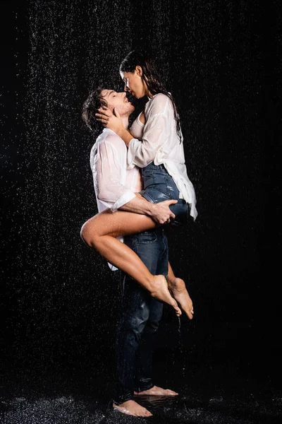 Сексуальная романтическая пара в мокрых белых рубашках и джинсах страстно обнимающаяся в дождевых капельках на черном фоне — стоковое фото
