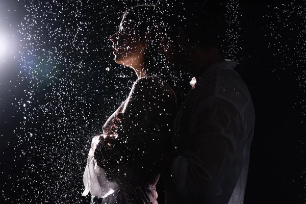 Sexy casal romântico apaixonadamente abraçando em gotas de chuva no fundo preto com luz traseira — Fotografia de Stock