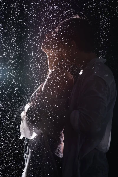 Sexy romantic couple passionately hugging in rain drops on black background with back light — Stock Photo