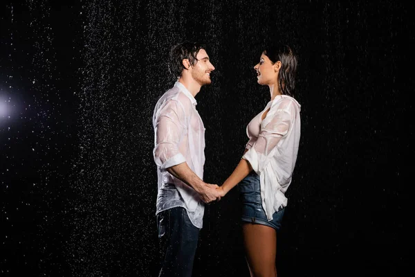 Sonriente pareja romántica en ropa mojada tomados de la mano y mirándose unos a otros en gotas de lluvia sobre fondo negro - foto de stock