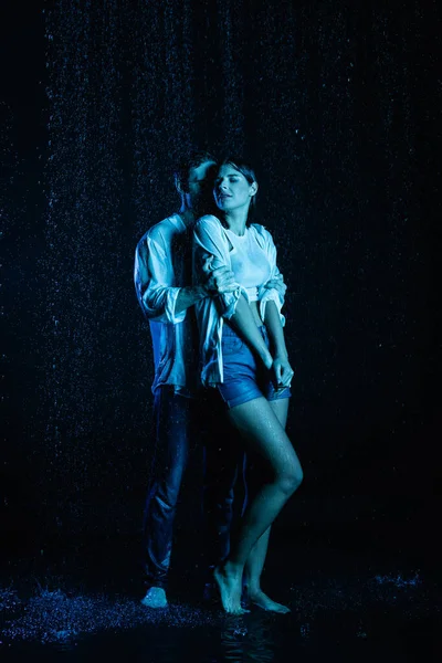 Пристрасний чоловік ніжно обіймає мокру чуттєву жінку в краплі води на чорному тлі з фільтром синього кольору — стокове фото