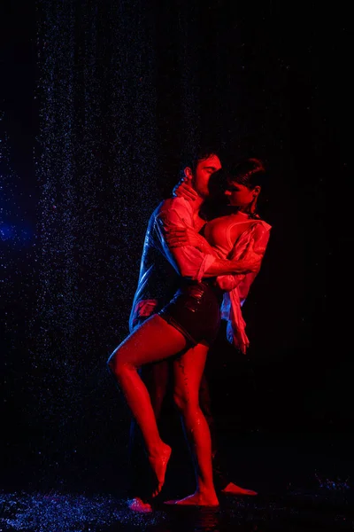 Wet passionate man hugging pretty woman in water drops, toned in red and blue on black background — Stock Photo