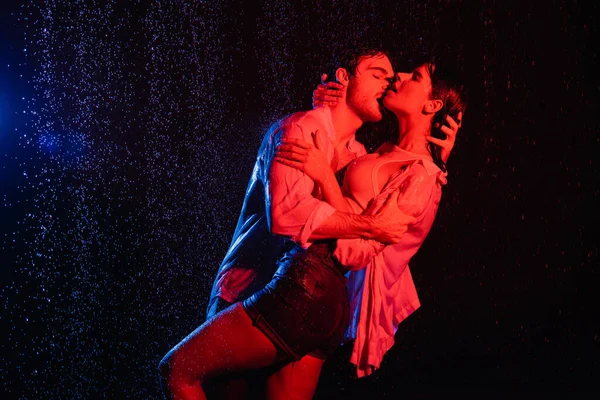 Humide sexy homme câlins et baisers jolie femme dans l'eau gouttes, tonique en rouge et bleu sur fond noir — Photo de stock