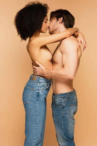 Вид збоку пристрасна міжрасова пара в джинсах тільки обіймає і цілує ізольовано на бежевому — стокове фото