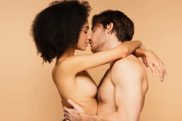 Вид збоку сексуальних багатоетнічних коханців, що обіймаються і цілуються із закритими очима ізольовані на бежевому — стокове фото
