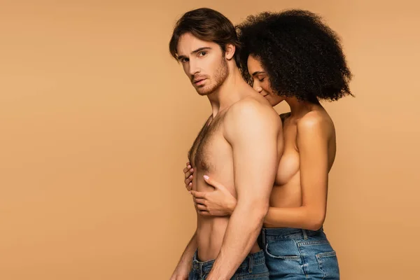 Topless mulher latina com olhos fechados abraçando tronco muscular de homem sem camisa olhando para a câmera isolada no bege — Fotografia de Stock